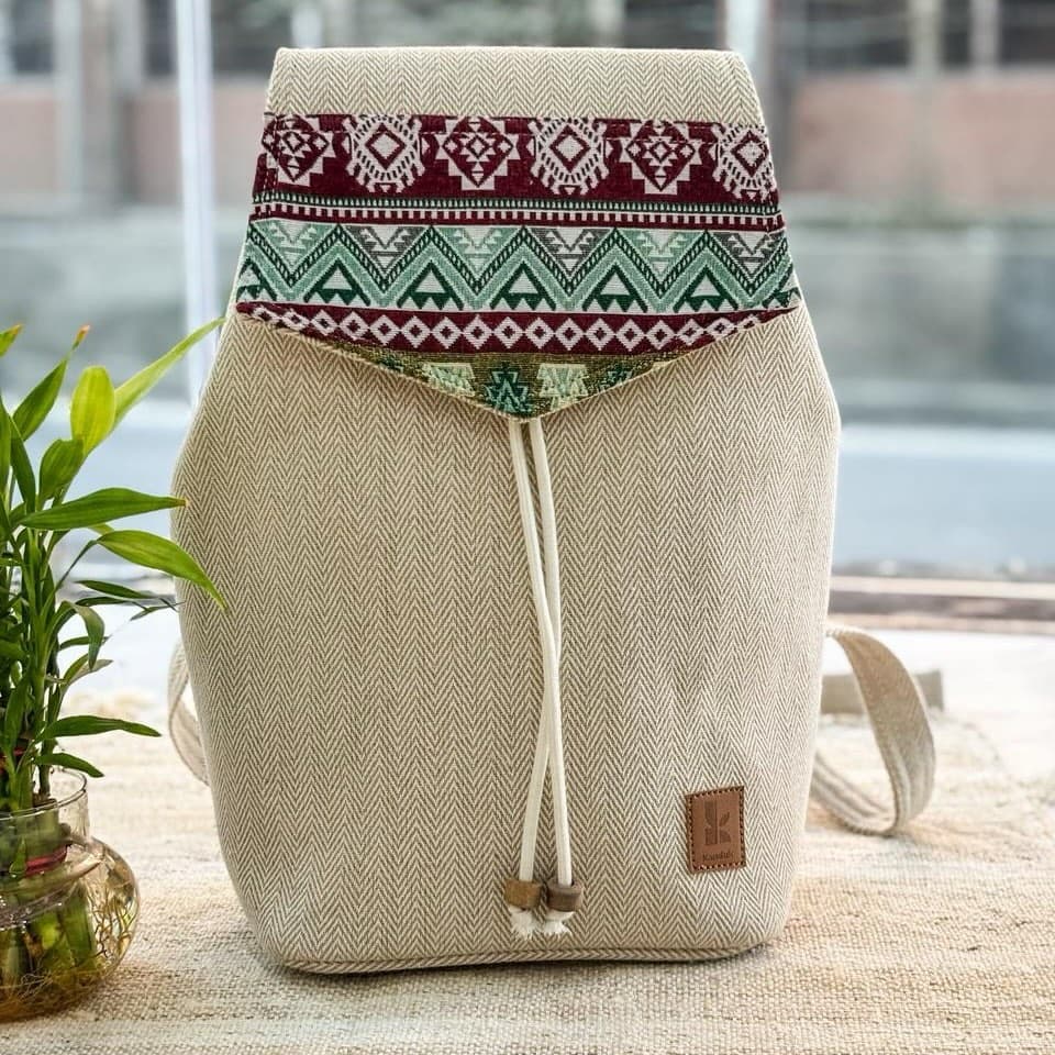 A natural herringbone hemp backpack with a crimson and green woven flap, standing on a wooden table beside a window