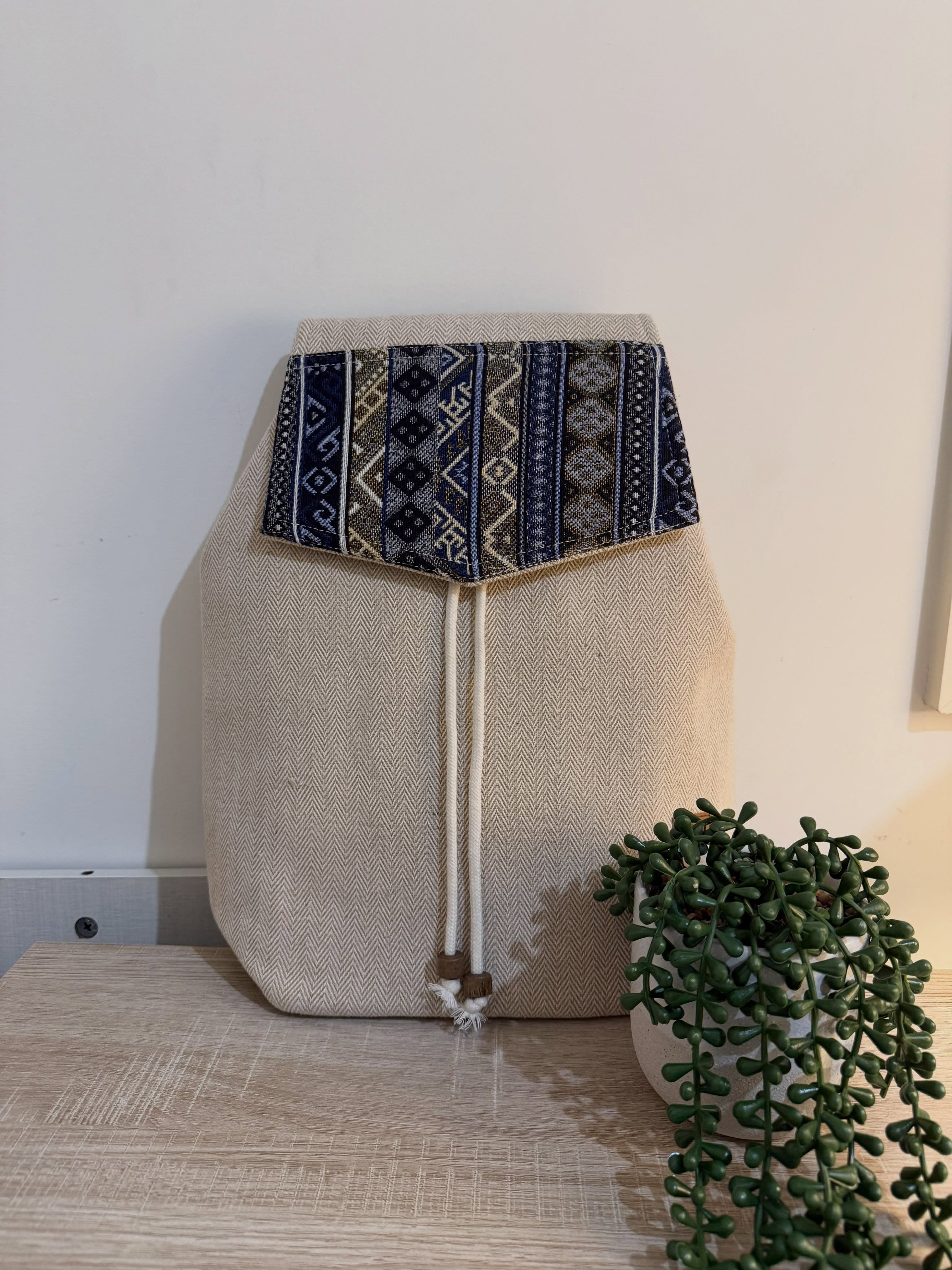 A natural herringbone hemp backpack with a navy and gold patterned flap, standing beside a potted plant