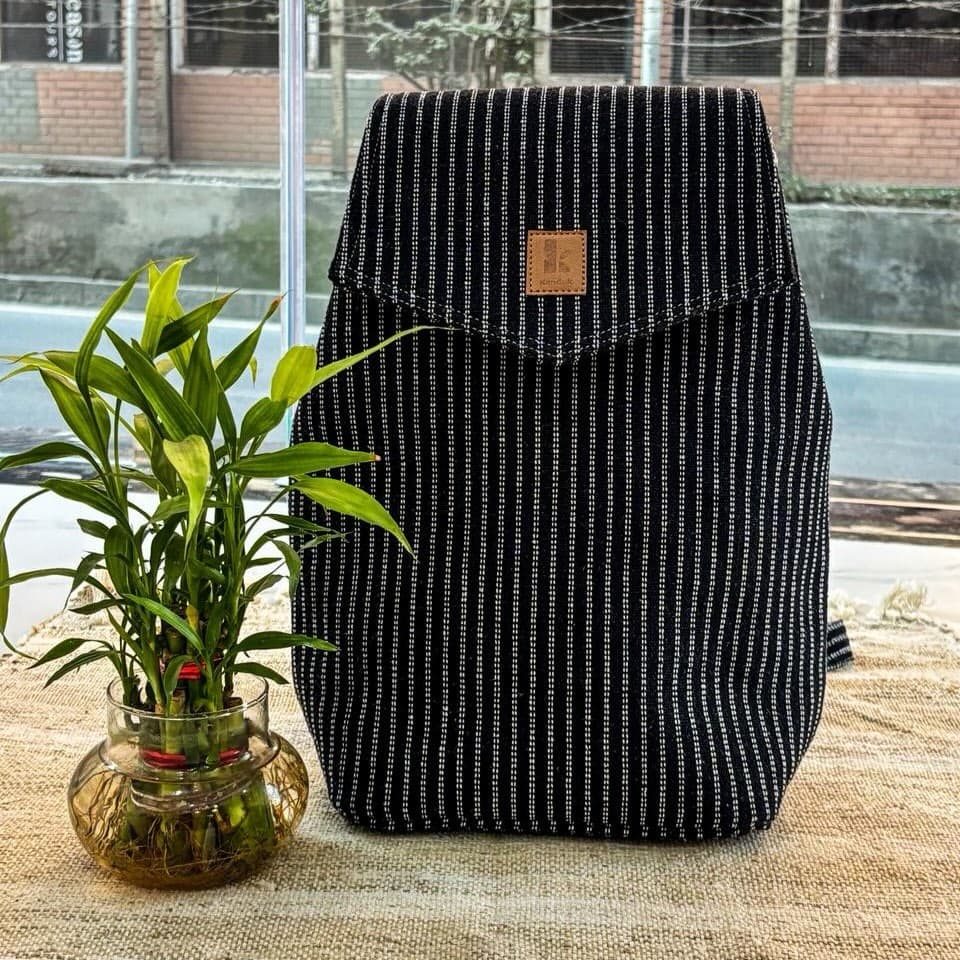 The Kanduk backpack in Midnight Pinstripe — a dark pinstriped hemp pack with a plain flap, standing on a table beside a bamboo plant